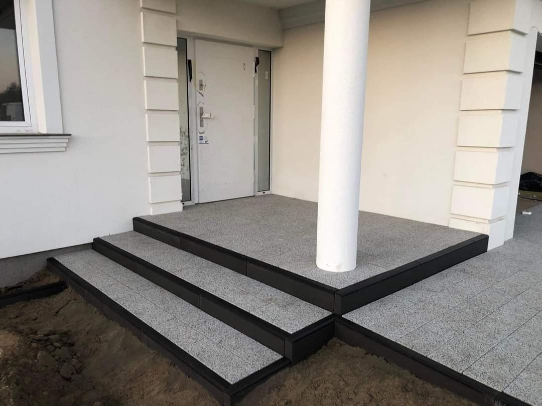 Nowoczesne schody wejściowe z szarej, drobnoziarnistej nawierzchni, obramowane czarnymi elementami, prowadzące do białych drzwi z kolumną przy wejściu.