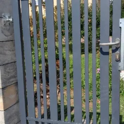 Szara furtka z pionowymi szczeblami, zamontowana na słupku z szarej kostki, z widocznym zamkiem i klamką, na tle zieleni ogrodu i domu z czerwoną dachówką.