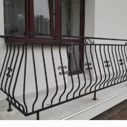 Czarna, metalowa balustrada balkonowa z dekoracyjnymi elementami w kształcie spirali, zamontowana na białej elewacji budynku z brązowym oknem.