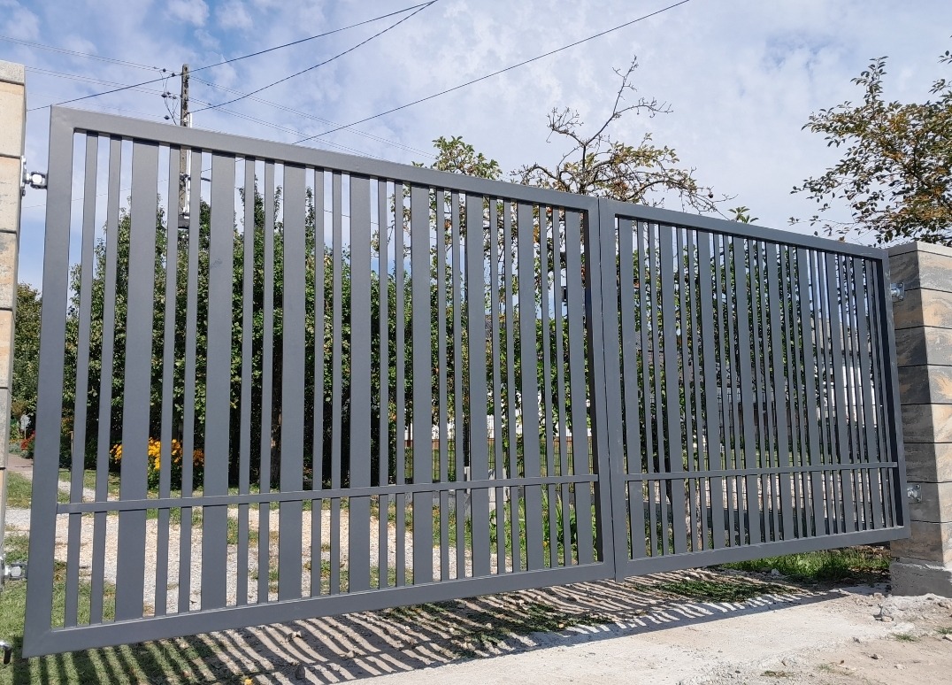 Nowoczesna, dwuskrzydłowa brama wjazdowa z pionowych, metalowych profili pomalowanych na szaro, zamontowana na słupkach z imitacji kamienia, widok z zewnątrz na tle zieleni.
