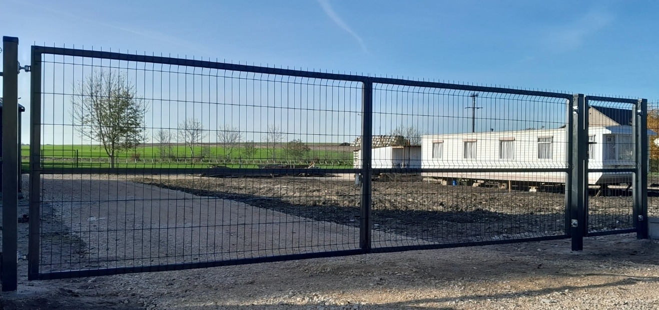Nowoczesna brama wjazdowa panelowa z metalową siatką na tle mobilnego domu i zielonego pola pod błękitnym niebem.