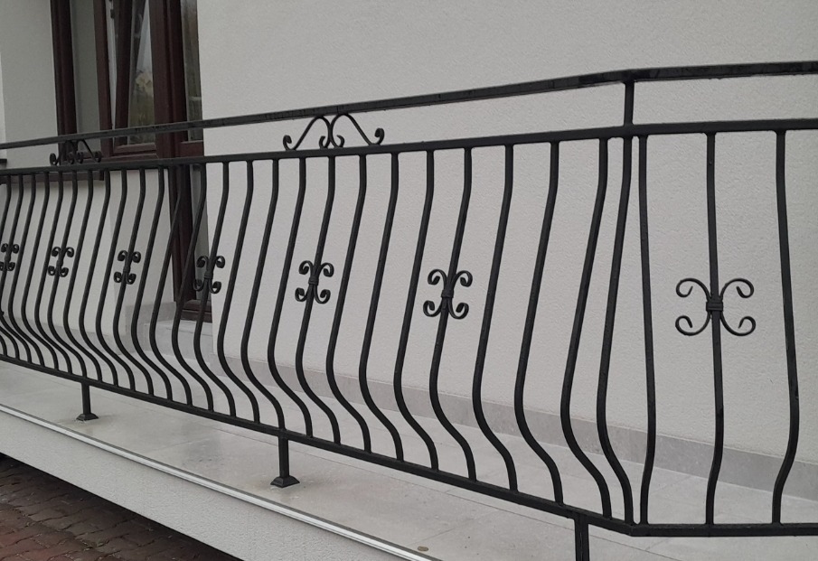Czarna, metalowa poręcz balkonowa z dekoracyjnymi, ażurowymi elementami w kształcie czterolistnej koniczyny, zamontowana na jasnym balkonie z widocznym fragmentem okna w tle.