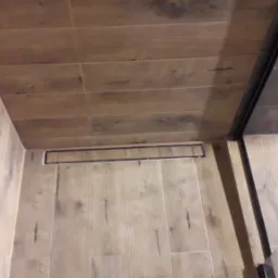 Nowoczesna kabina prysznicowa z odpływem liniowym, wykończona płytkami imitującymi drewno na ścianach i podłodze, z czarnymi elementami armatury i szklaną ścianką.