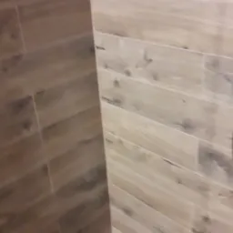 Fragment wykończonej ściany i podłogi w łazience, wyłożonych płytkami imitującymi drewno w odcieniach beżu i brązu, z widocznym narożnikiem.