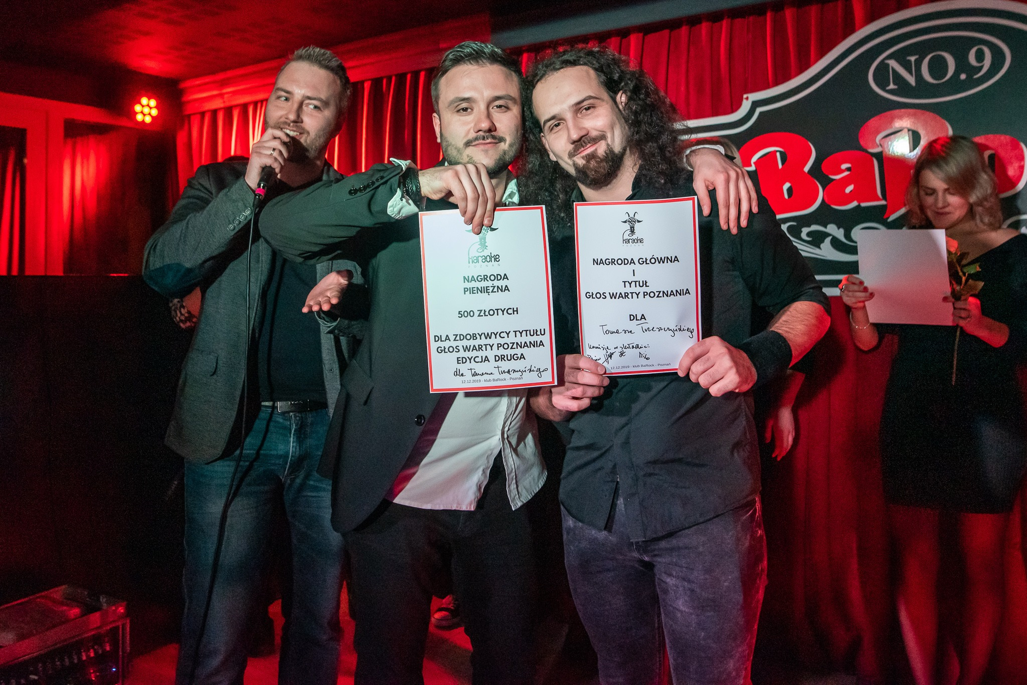 Zespół muzyczny odbiera nagrody pieniężne i tytuł Głosu Wartego Poznania podczas występu w klubie BaRock, trzymając dyplomy na tle czerwonej sceny.