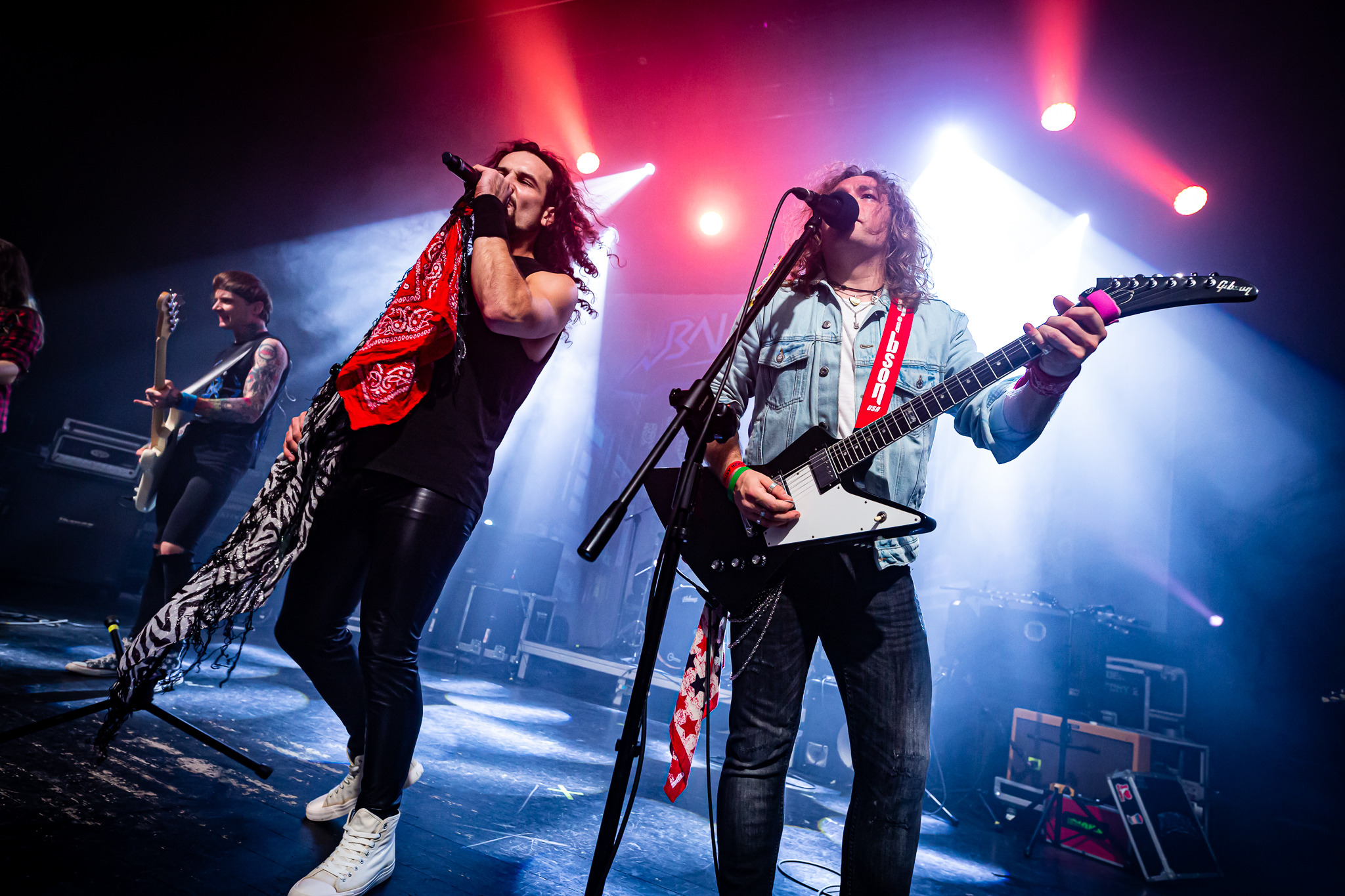 Dynamiczne ujęcie zespołu rockowego podczas występu na scenie w Poznaniu, z wokalistą w czerwonej bandanie i gitarzystą w dżinsowej kurtce, oświetlonych reflektorami.