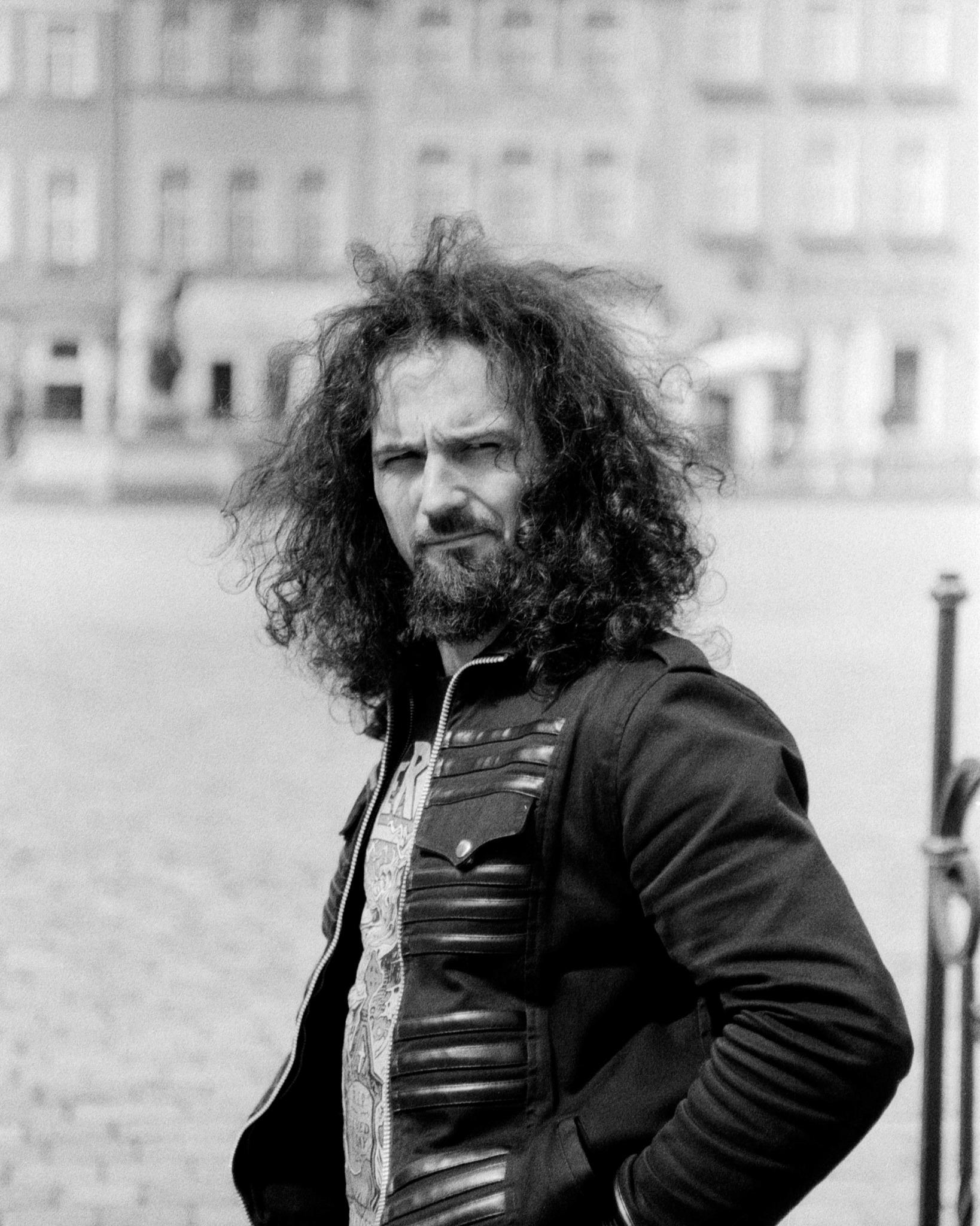 Czarno-biały portret muzyka z kręconymi włosami i brodą, w rozpiętej kurtce, na tle miejskiego placu z budynkami.