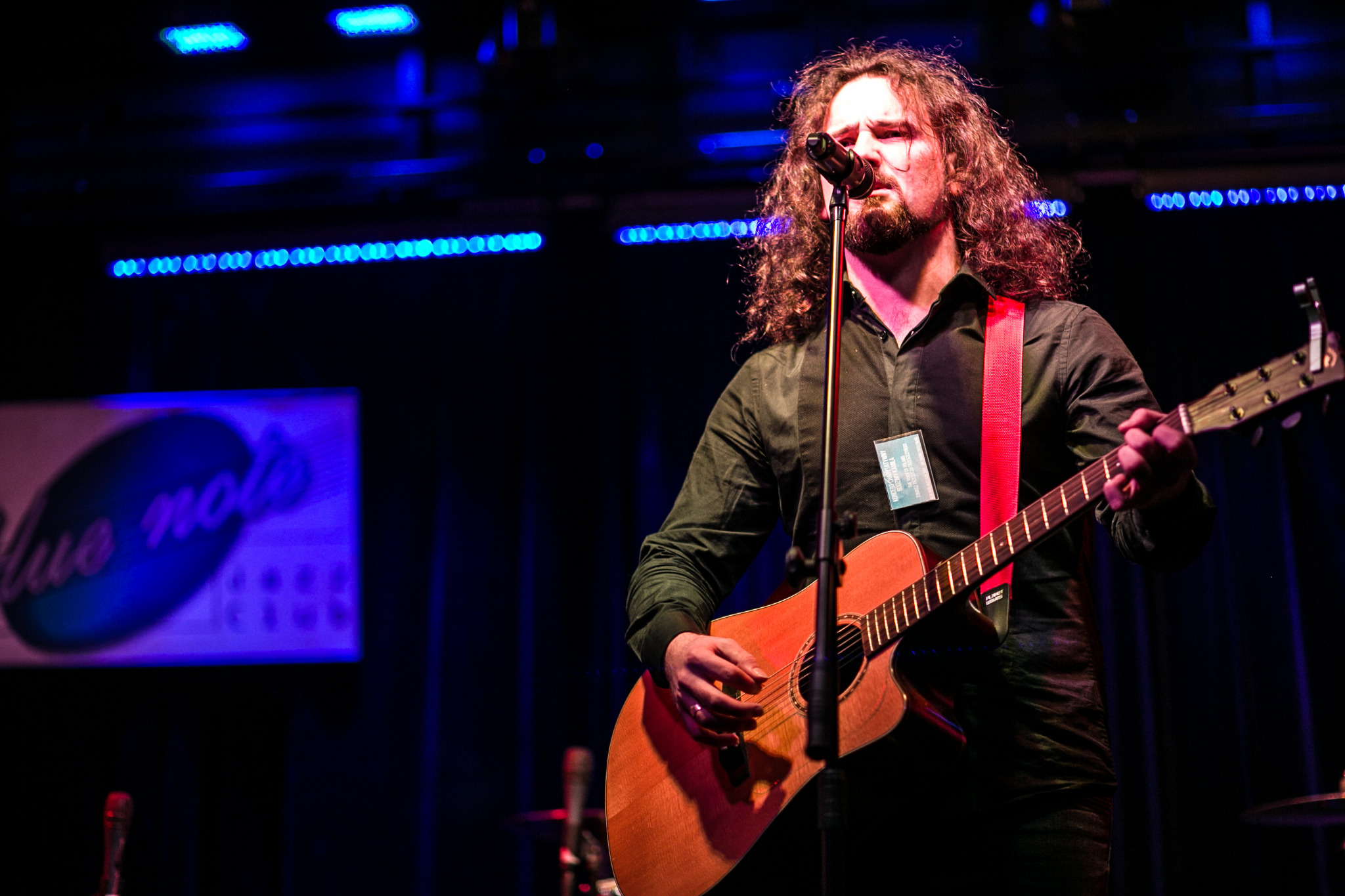 Wokalista z długimi kręconymi włosami śpiewa do mikrofonu grając na akustycznej gitarze na scenie z niebieskim oświetleniem.