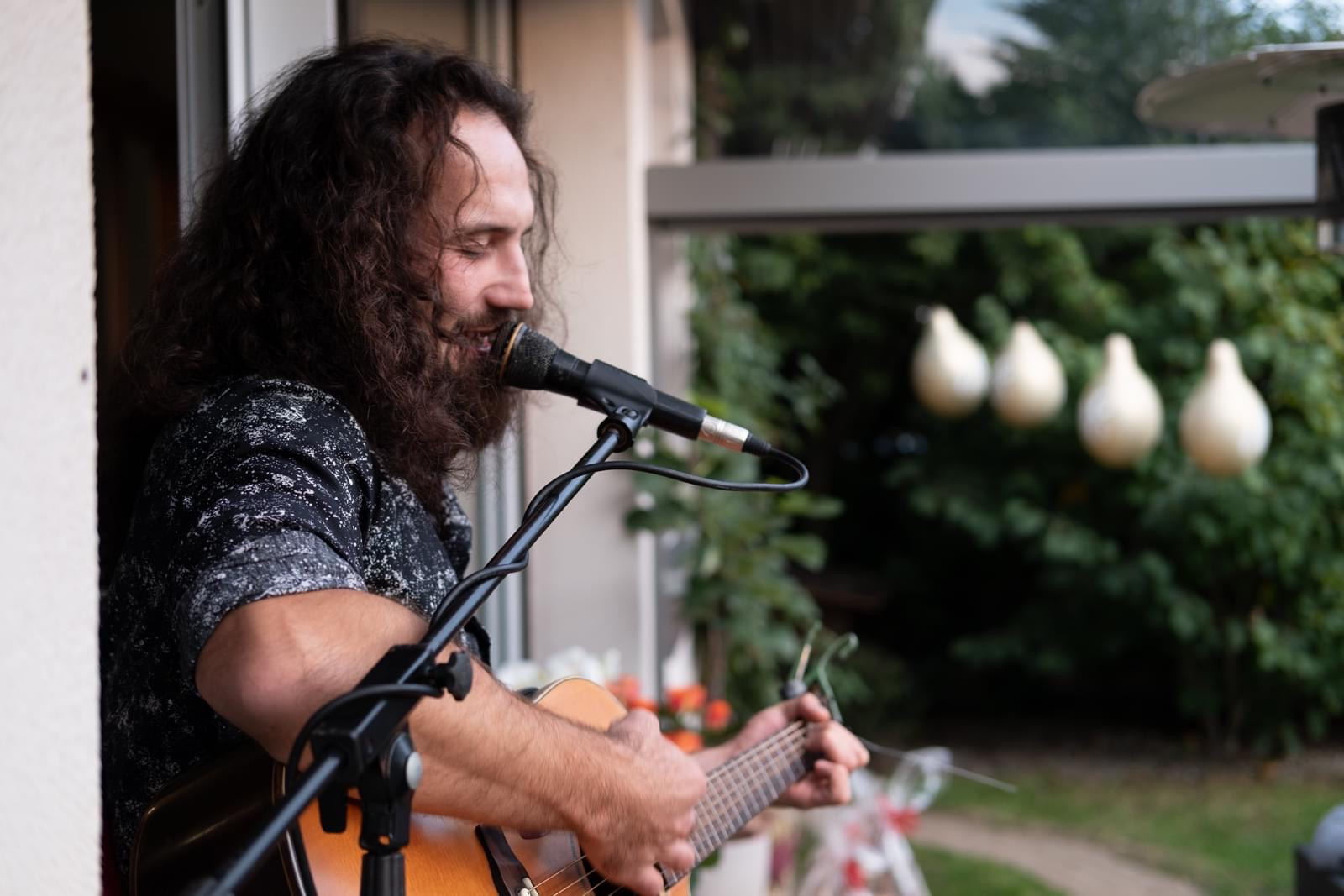 Muzyk z długimi kręconymi włosami i brodą gra na gitarze akustycznej i śpiewa do mikrofonu na zewnątrz, w tle widoczne dekoracje ogrodowe.