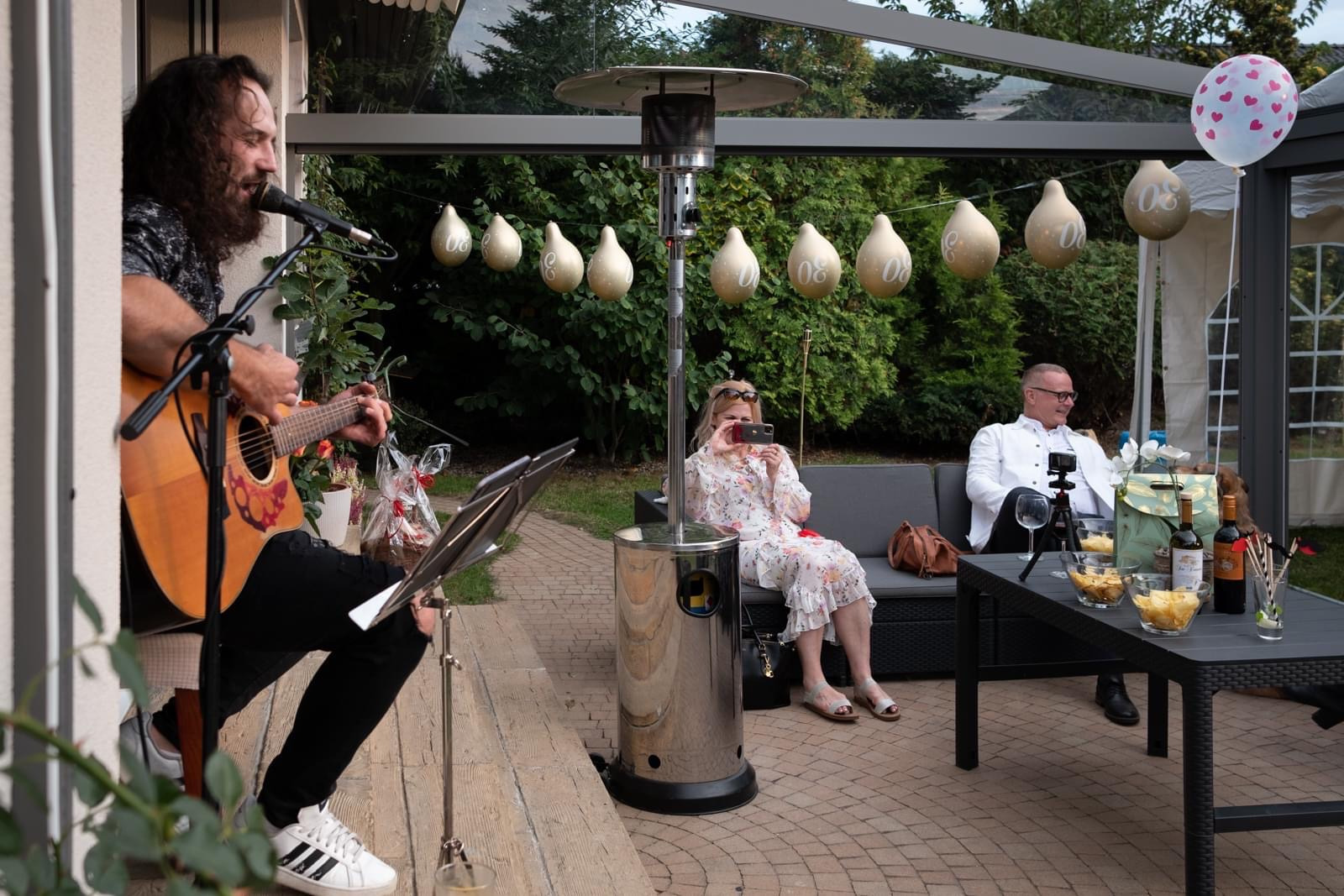 Mężczyzna z długimi włosami gra na gitarze akustycznej i śpiewa do mikrofonu podczas przyjęcia na zewnątrz; w tle goście siedzą na sofie, a dekoracje obejmują balony z liczbą 30 i grzejnik tarasowy.