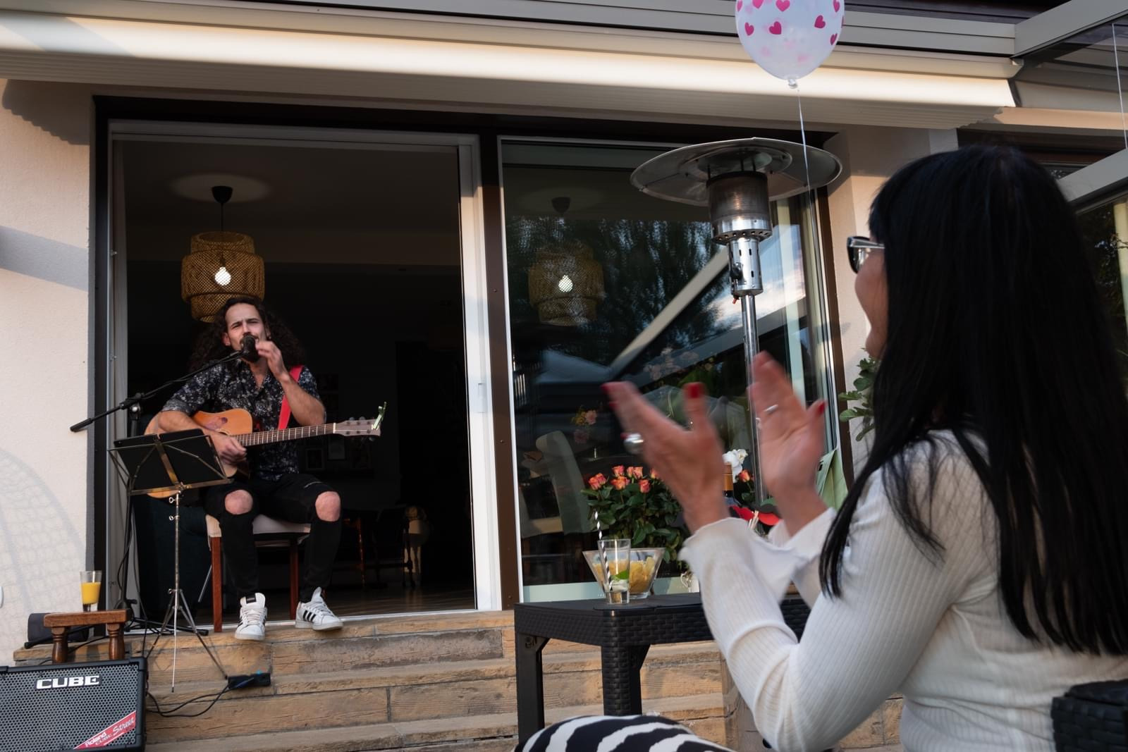 Muzyk z długimi włosami gra na gitarze akustycznej na schodach, ubrany w podarte jeansy i wzorzystą koszulę, z mikrofonem i wzmacniaczem Cube; kobieta w białej bluzce klaszcze, na stoliku drinki...