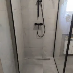 Nowoczesna kabina prysznicowa typu walk-in z czarną armaturą i odpływem liniowym w podłodze, wykończona płytkami imitującymi marmur, zabezpieczona żółtą taśmą malarską na łączeniach szkła.