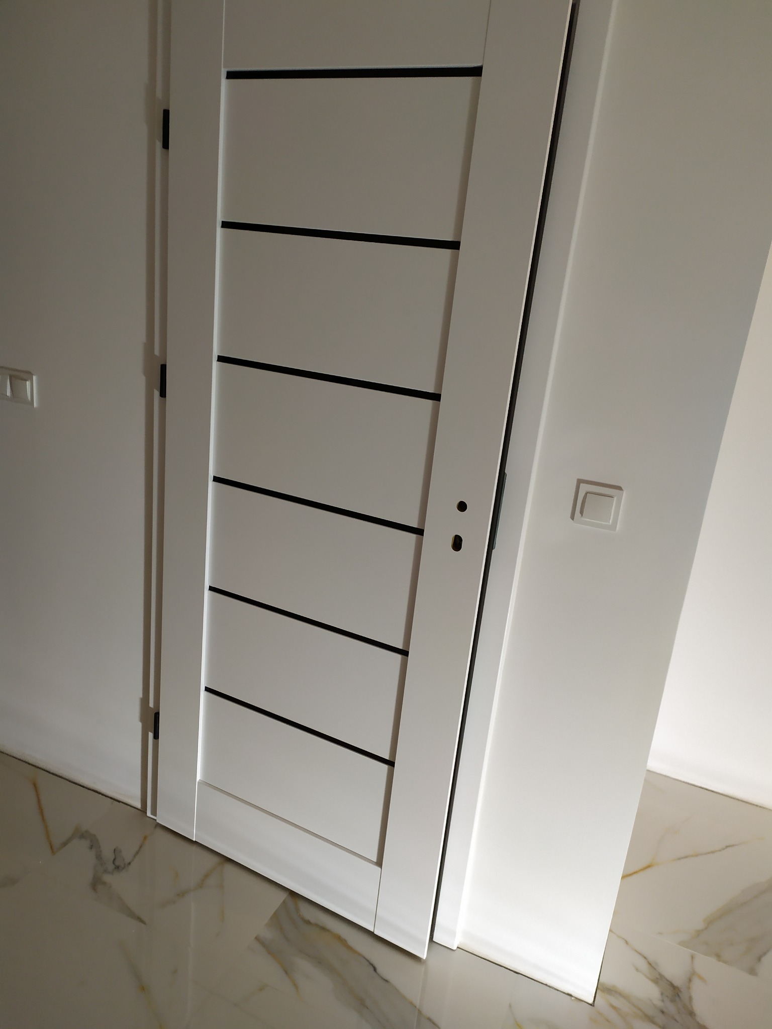 Nowoczesne, białe drzwi wewnętrzne z czarnymi, poziomymi akcentami, zamontowane w białej ścianie z widocznym włącznikiem światła i marmurową podłogą.