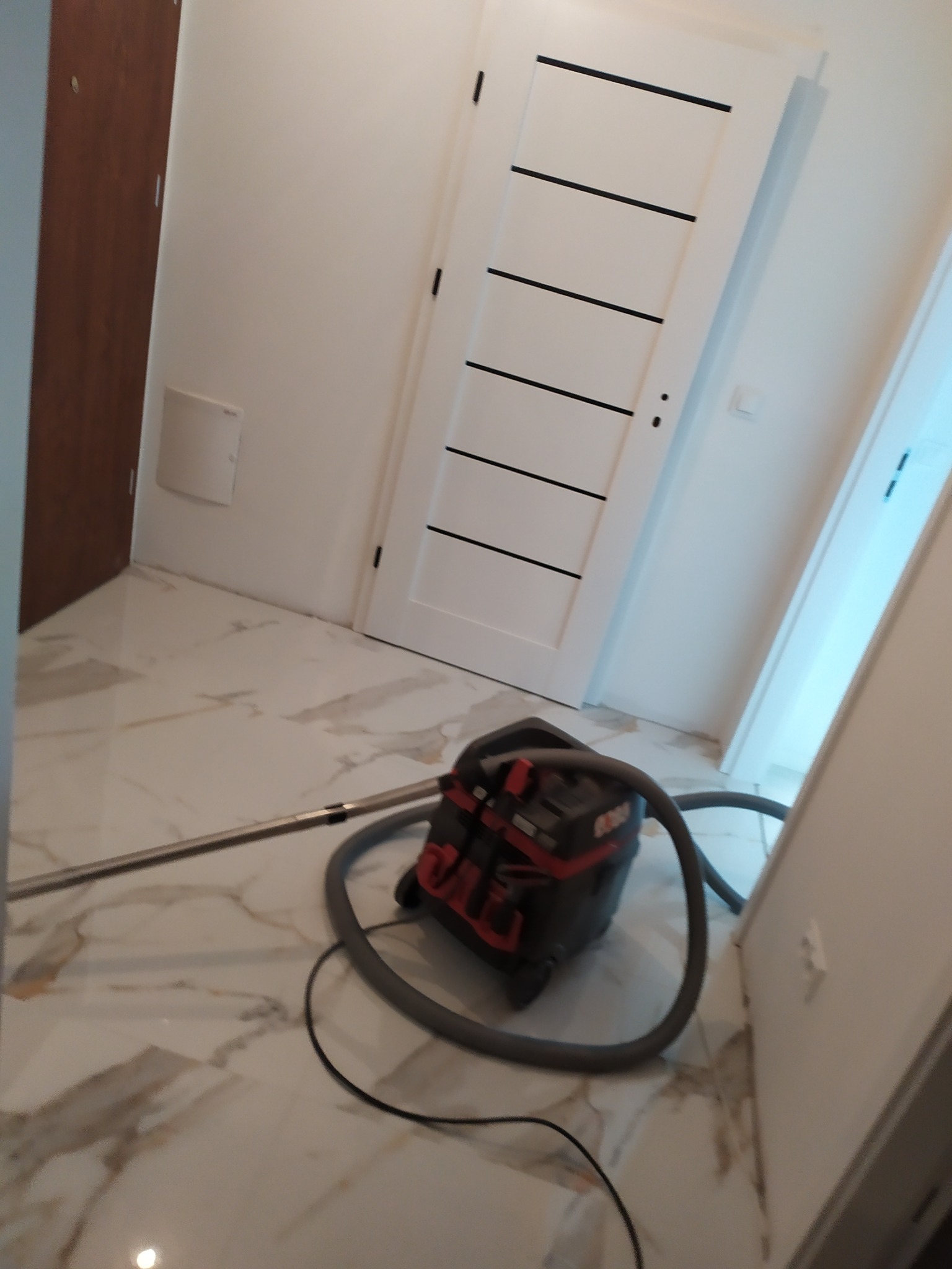 Wnętrze po remoncie z marmurową podłogą, białe drzwi z poziomymi czarnymi paskami i odkurzacz przemysłowy na środku, sugerujący prace porządkowe po wykonanej usłudze.