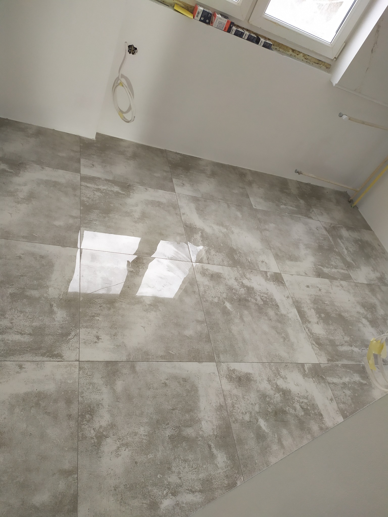 Ściana wyłożona szarymi, matowymi płytkami imitującymi beton, widoczne gniazdko elektryczne i fragment instalacji pod sufitem, okno dachowe w tle.