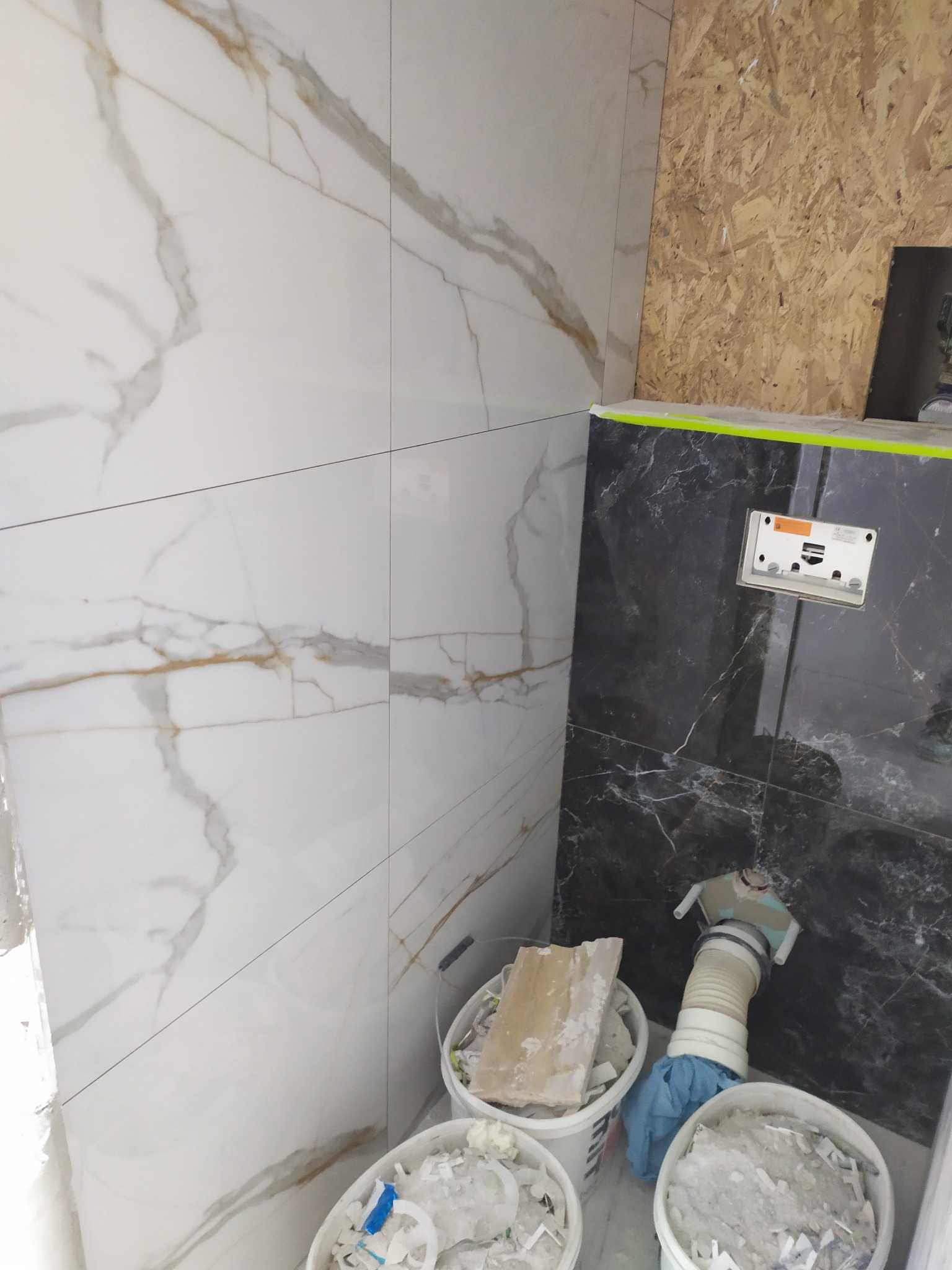 Ściana łazienki wyłożona białymi i czarnymi płytkami imitującymi marmur, z widocznym fragmentem instalacji i wiadrami z resztkami materiałów budowlanych.