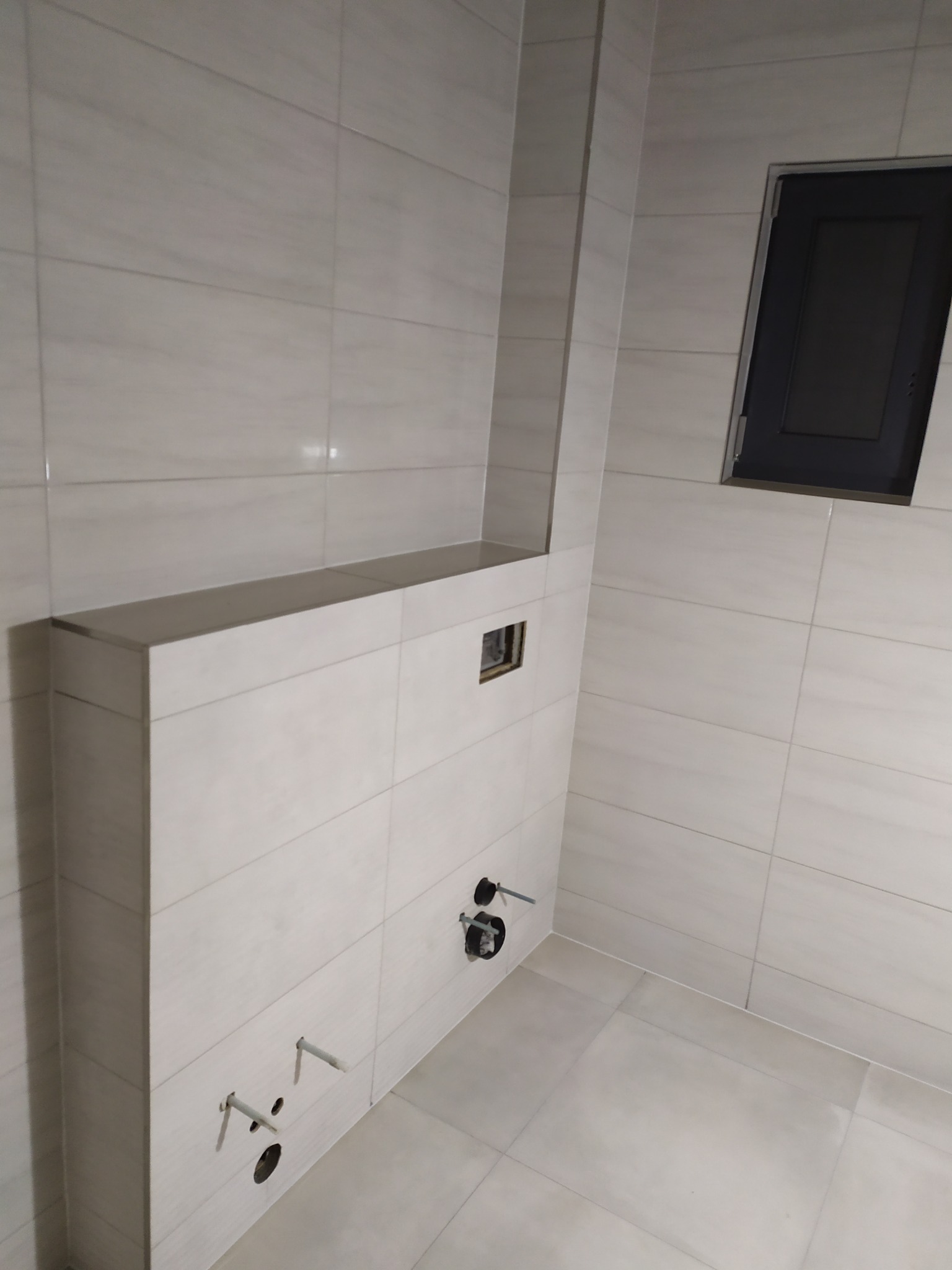 Wnętrze łazienki w trakcie prac: obudowa stelaża WC z płyt gipsowo-kartonowych, widoczne przyłącza wodne, szara wentylacja i jasne płytki ceramiczne na ścianach i podłodze.