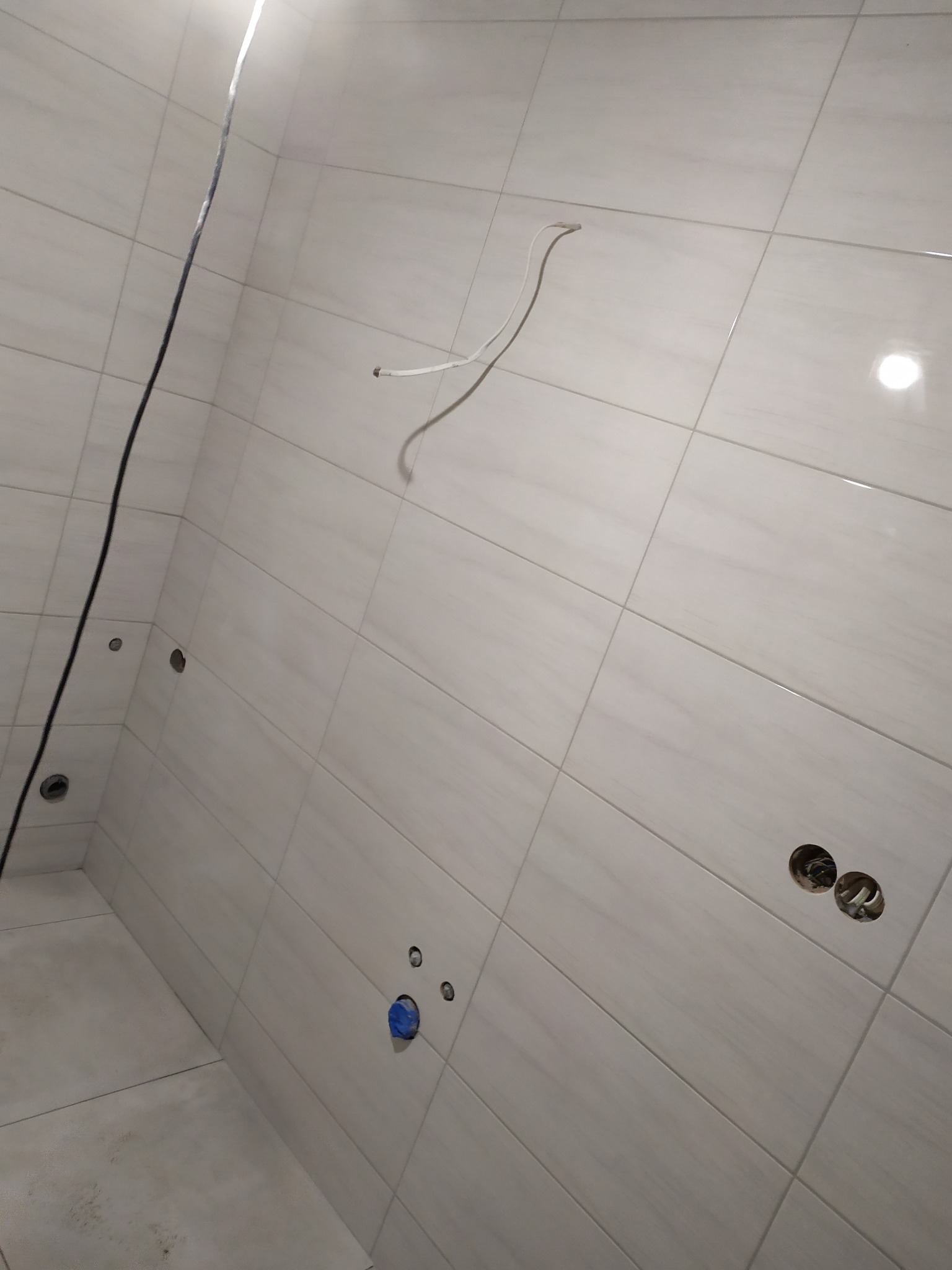 Ściana i podłoga w trakcie kładzenia płytek w łazience, widoczne kable elektryczne i rury wodne przygotowane do podłączenia armatury.