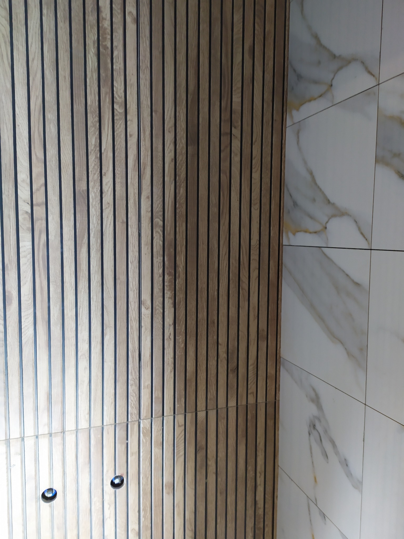 Fragment łazienki z drewnianą ścianą z lameli i marmurowymi płytkami. Widoczne dwa punkty świetlne w suficie.