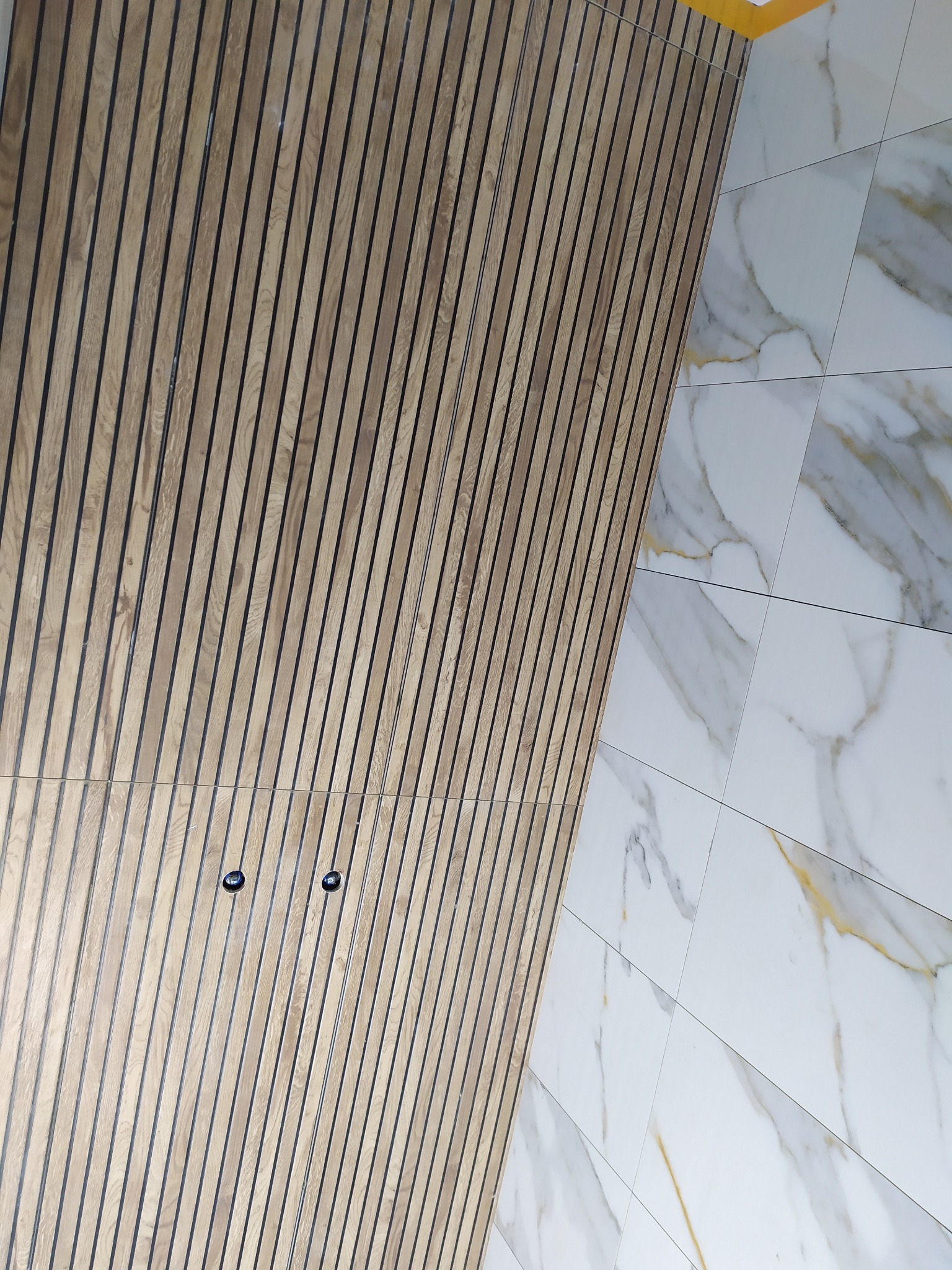 Ściana wykończona panelami drewnianymi z czarnymi przerwami, sąsiadująca z powierzchnią z płytek imitujących marmur z złotymi żyłami, widoczne otwory na gniazdka elektryczne.