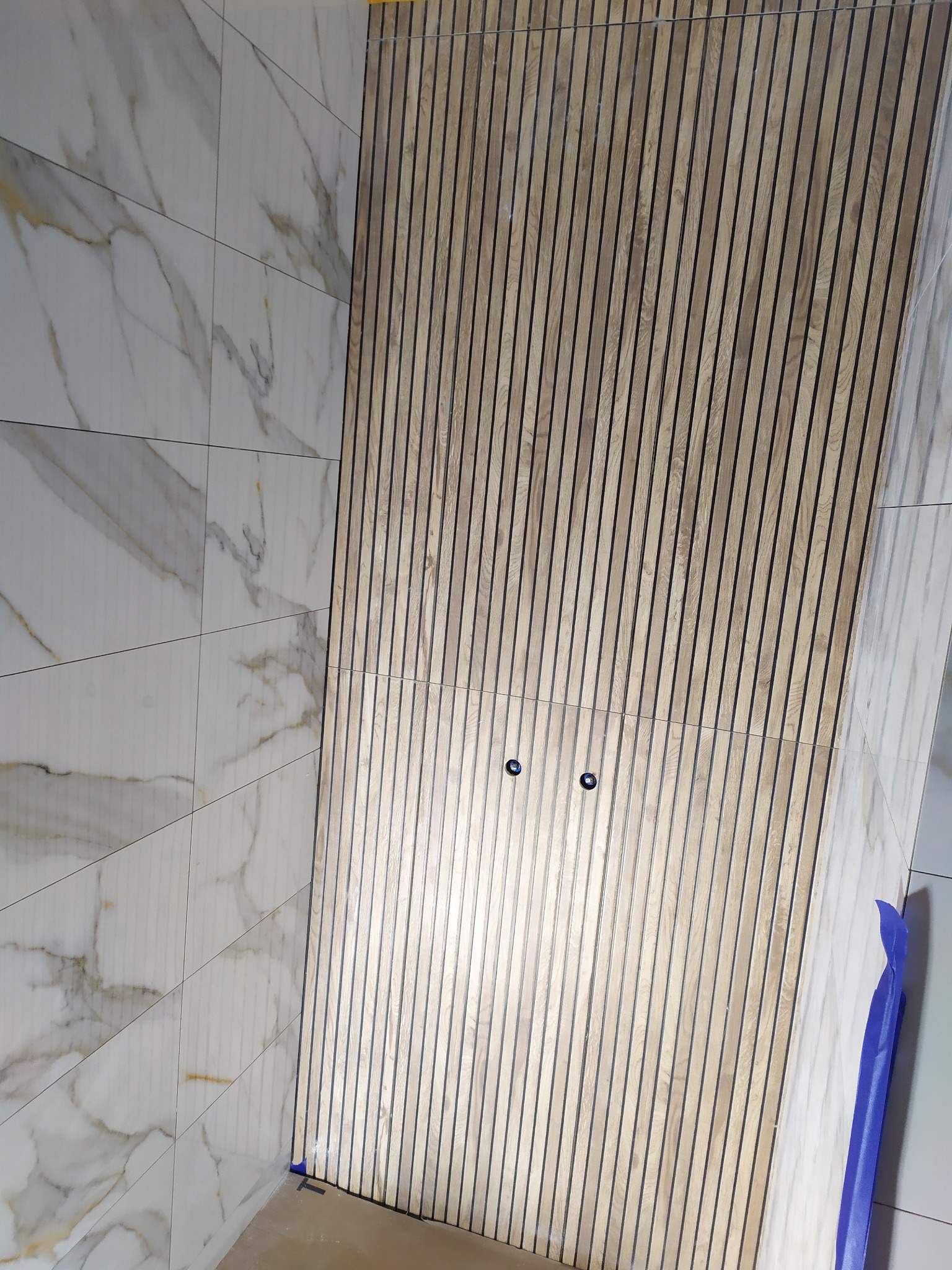 Fragment ściany w łazience z panelami drewnianymi i płytkami imitującymi marmur, widoczne dwa otwory na oświetlenie, zabezpieczona folią krawędź.