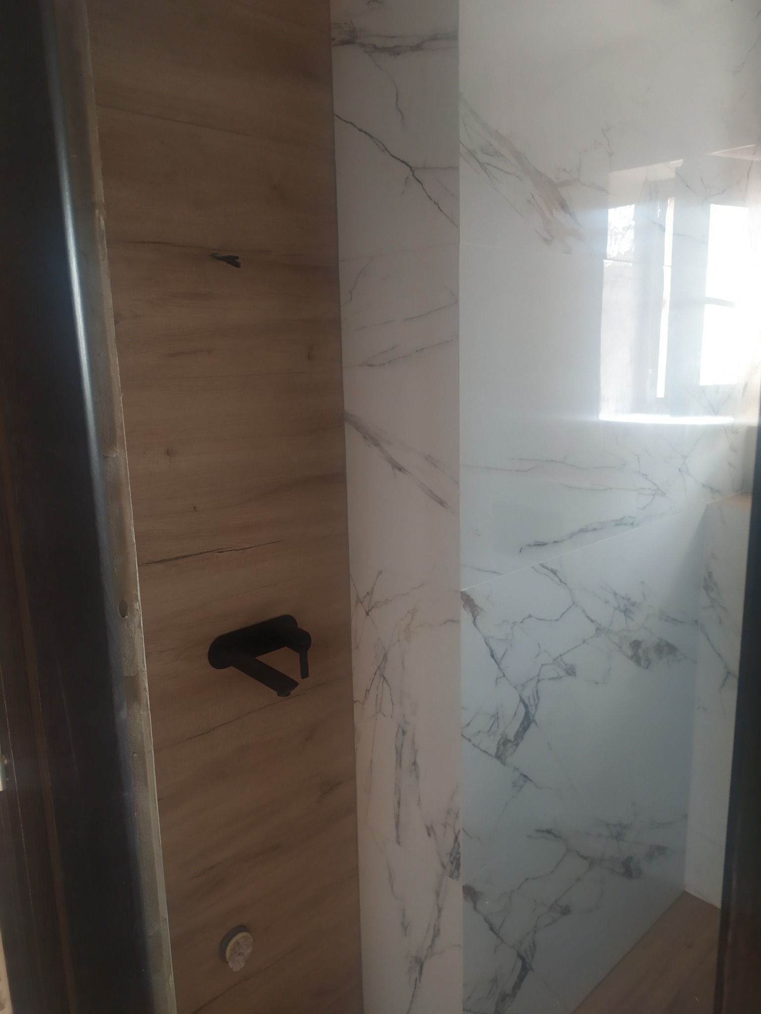 Fragment wykończonej łazienki: drewnopodobna płyta ścienna z czarną armaturą i białe płytki imitujące marmur na ścianie prysznica.