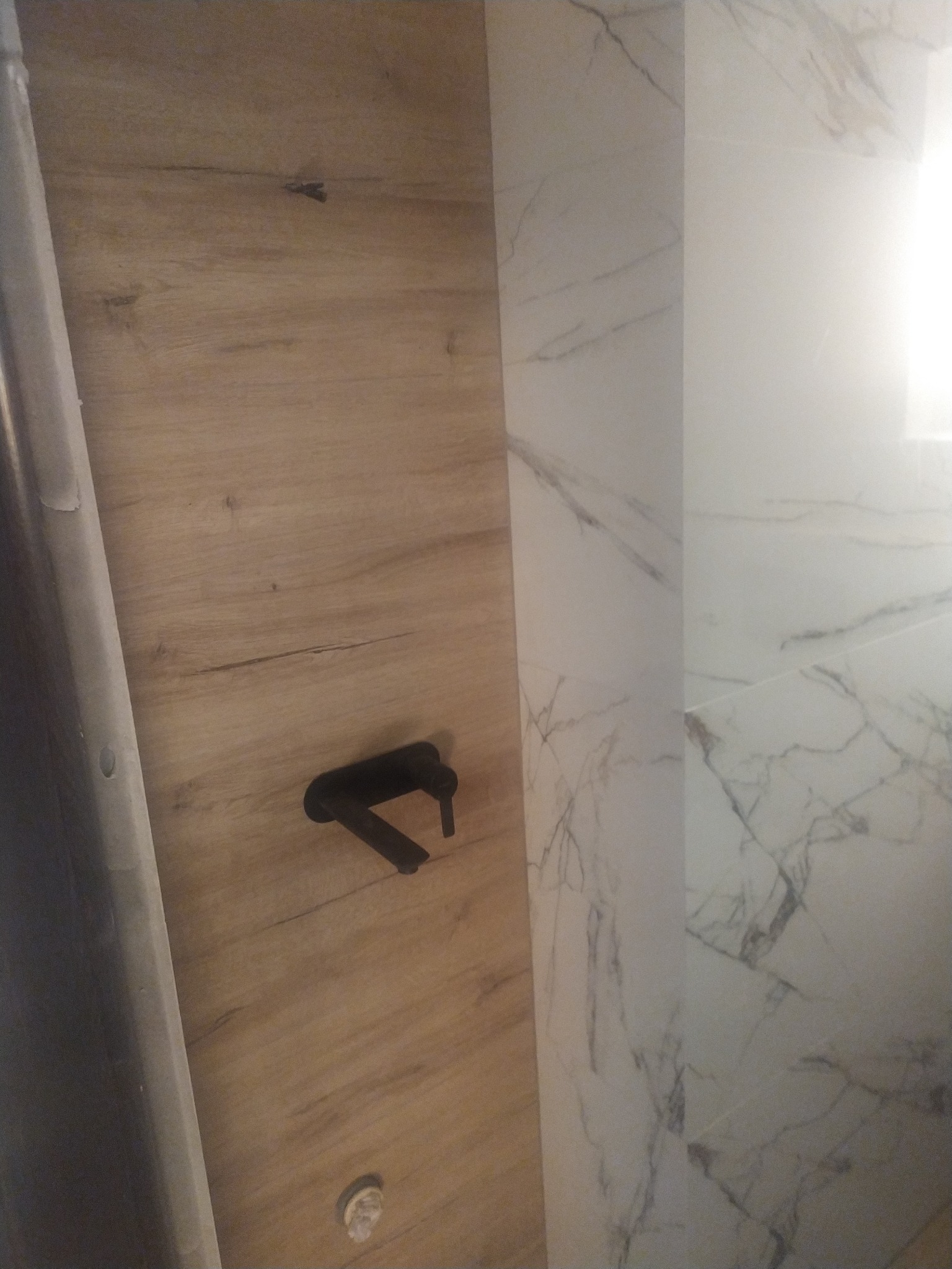 Fragment łazienki z drewnopodobną okładziną ścienną, czarna bateria umywalkowa i marmurowe płytki na ścianie obok.