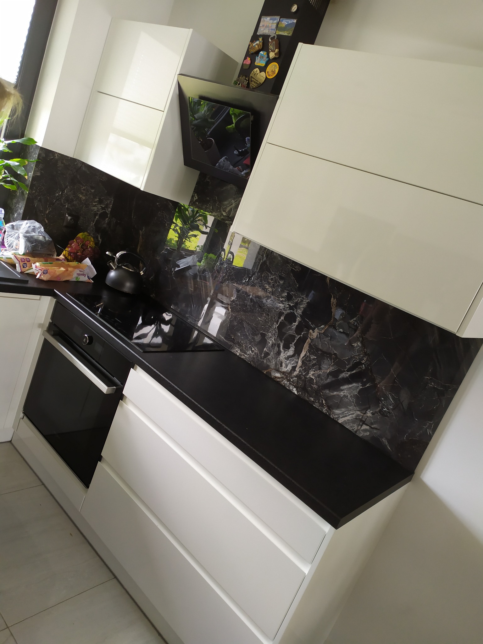 Nowoczesna kuchnia z białymi szafkami, czarnym blatem i marmurową ścianą nad blatem, na blacie czajnik i produkty spożywcze.