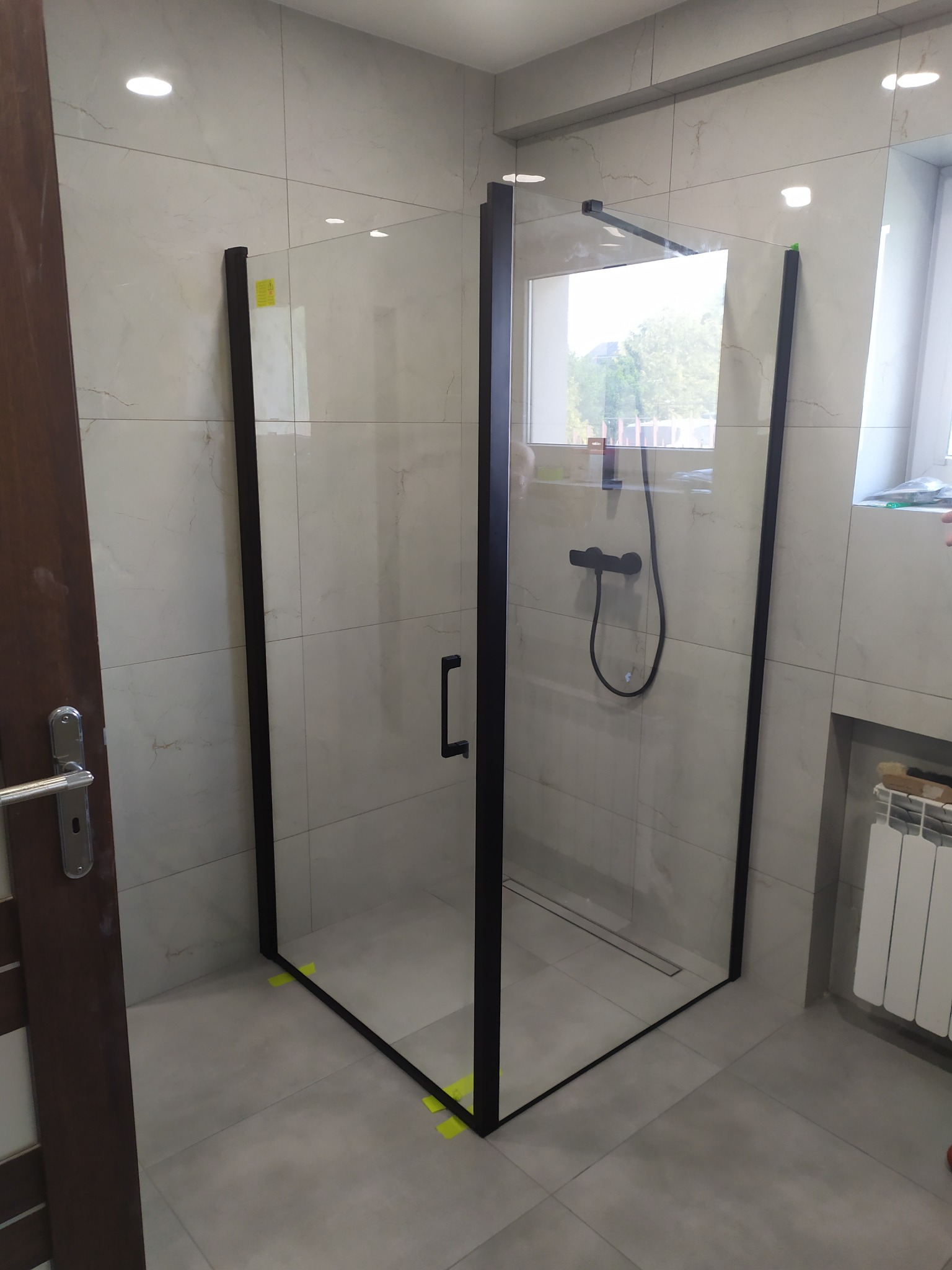 Nowoczesna kabina prysznicowa z czarnymi profilami i deszczownicą, zamontowana w łazience wykończonej płytkami imitującymi marmur, z widocznymi żółtymi zabezpieczeniami montażowymi.