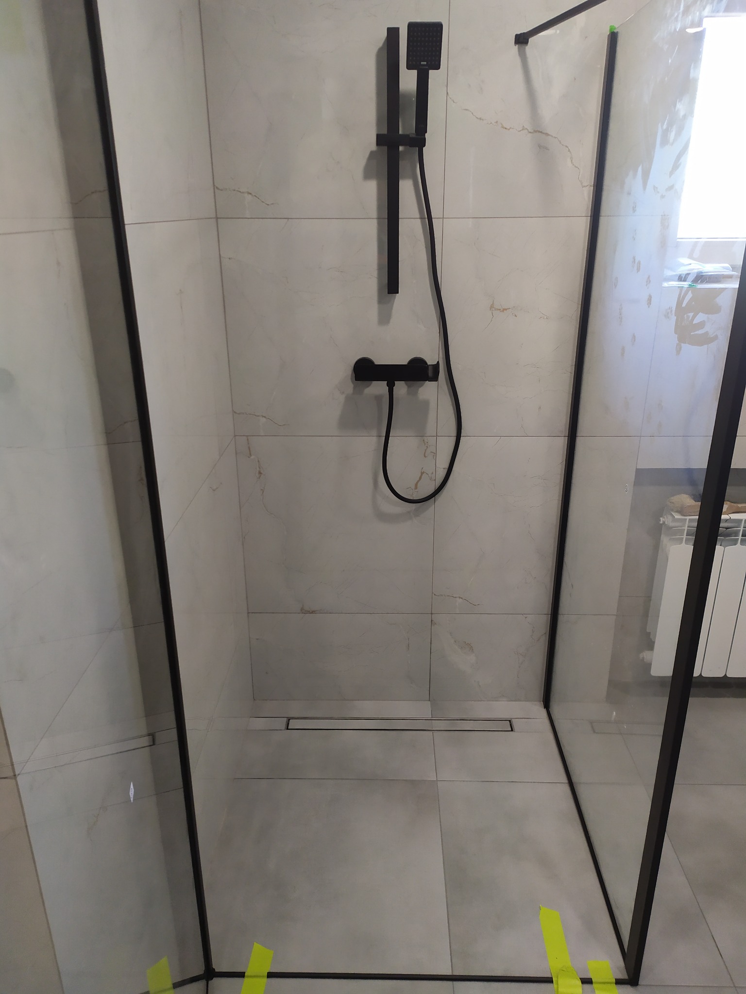 Nowoczesna kabina prysznicowa typu walk-in z czarną armaturą i odpływem liniowym w podłodze, wykończona płytkami imitującymi marmur, zabezpieczona żółtą taśmą malarską na łączeniach szkła.