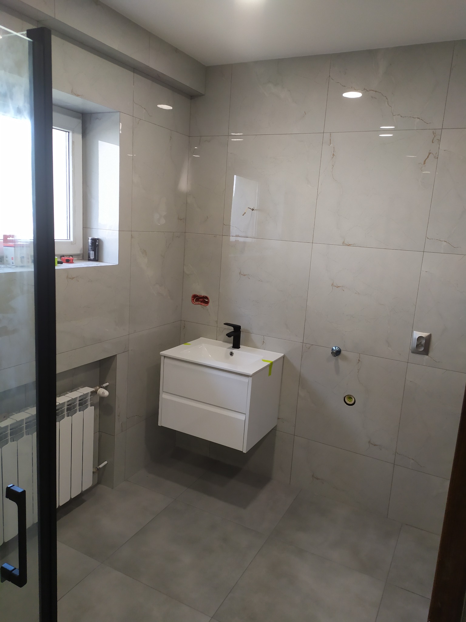 Nowoczesna łazienka z marmurowymi płytkami, białą szafką pod umywalkę z czarną armaturą, grzejnikiem drabinkowym i czarną ramą kabiny prysznicowej, widoczne puszki instalacyjne w ścianie.