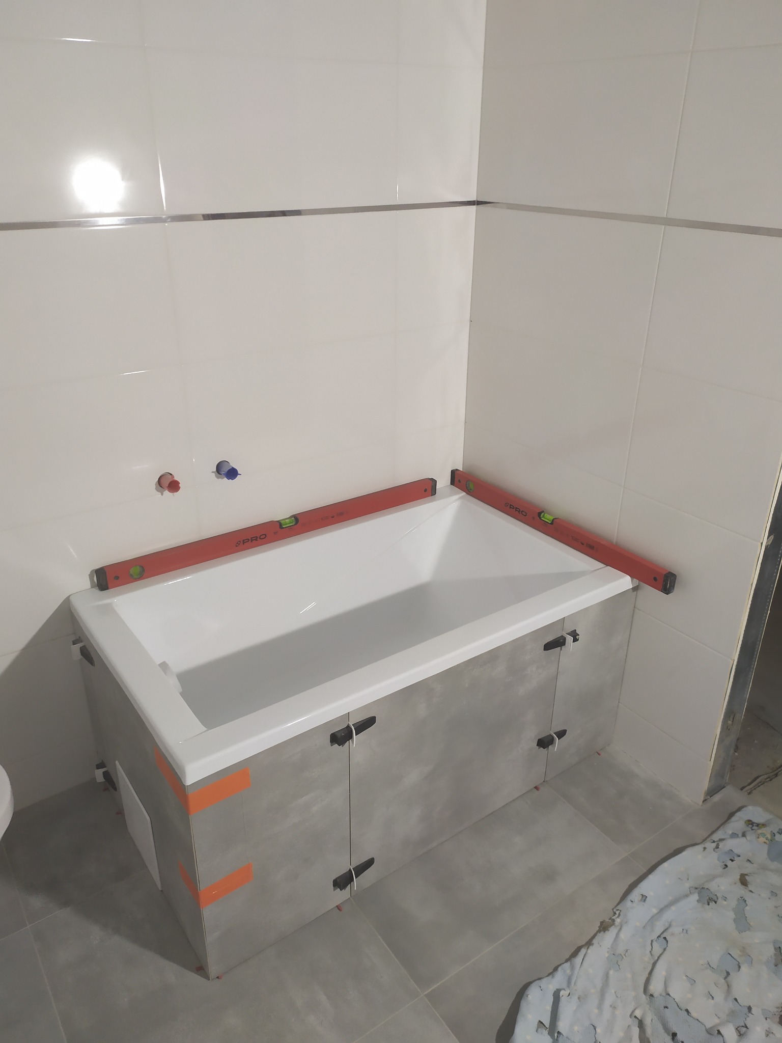 Biała wanna obudowana szarymi płytkami, zabezpieczonymi pomarańczową taśmą, poziomowana dwoma poziomicami PRO, z widocznymi przyłączami wodnymi na ścianie z białych płytek z metalowym paskiem...