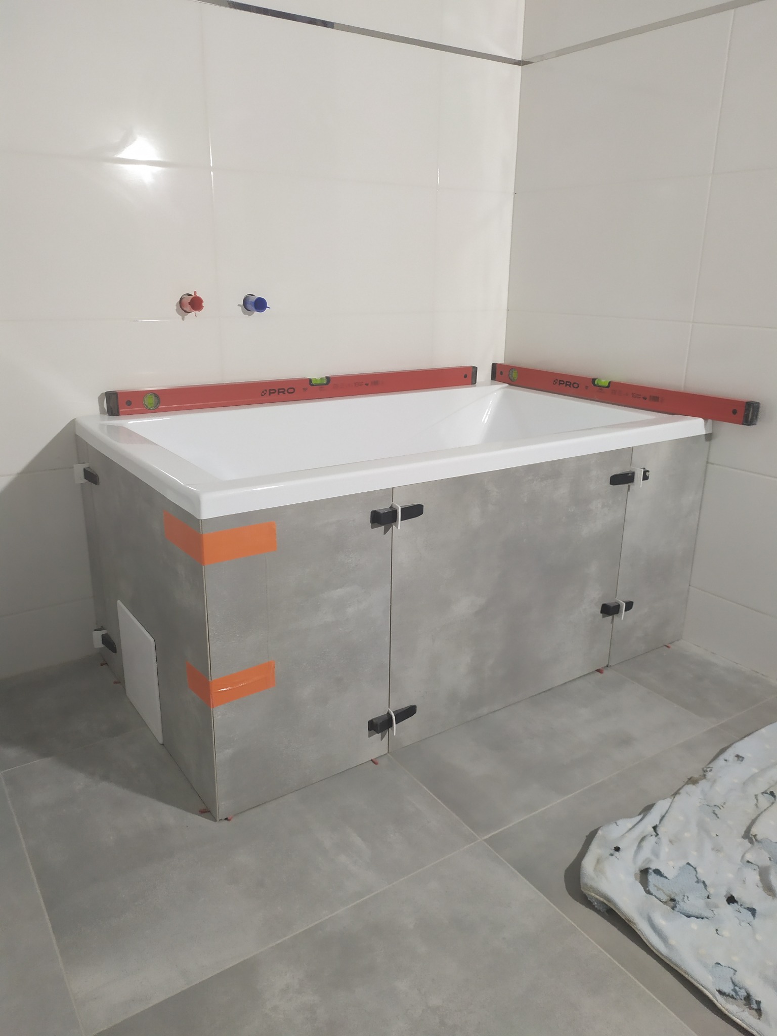 Obudowa wanny z szarych płytek imitujących beton w trakcie montażu, poziomice PRO na krawędziach, pomarańczowa taśma maskująca, białe płytki na ścianach, szare płytki podłogowe, widoczne krzyżyki...