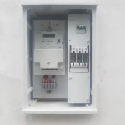 Otwarta skrzynka elektryczna z zamontowanym licznikiem NORAX 3, bezpiecznikami i listwą zaciskową, umieszczona na białej ścianie.