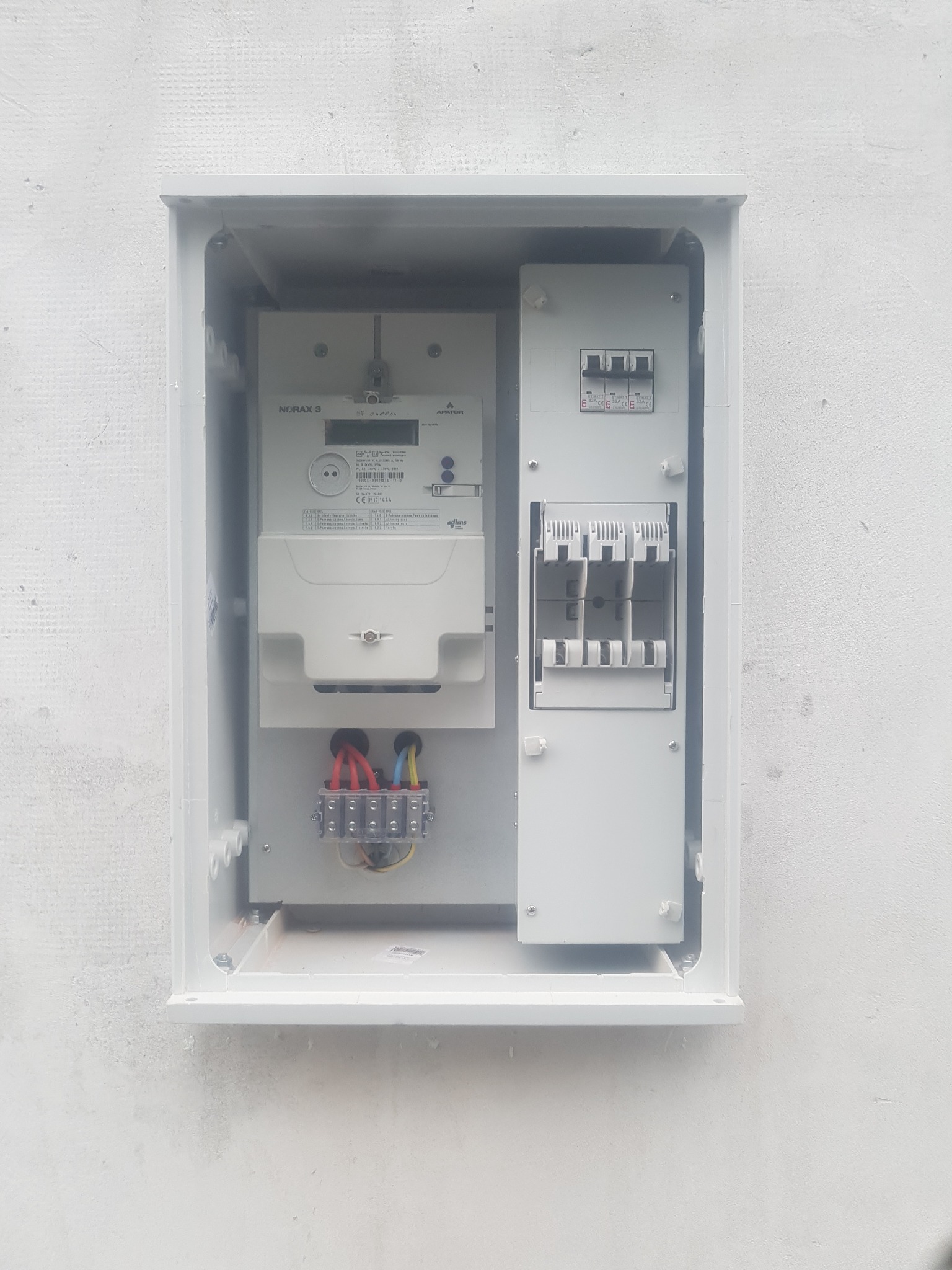 Otwarta skrzynka elektryczna z zamontowanym licznikiem NORAX 3, bezpiecznikami i listwą zaciskową, umieszczona na białej ścianie.