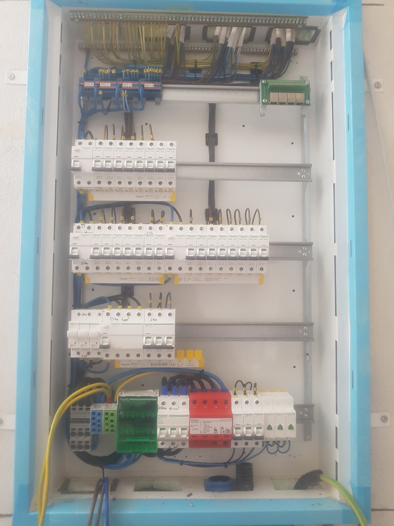 Otwarta skrzynka elektryczna z zainstalowanymi bezpiecznikami, przewodami i listwami zaciskowymi. Widoczne przewody w kolorach żółtym, niebieskim i czarnym. Elementy oznaczone RCD L1, L2, L3.