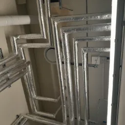 Srebrne, izolowane rury wentylacyjne poprowadzone pod sufitem, widoczne elementy mocujące i oświetlenie, ujęcie z dołu.