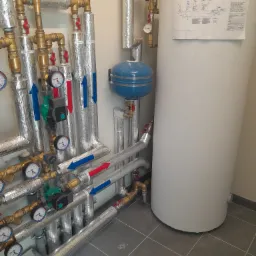 Szczegółowe ujęcie instalacji grzewczej z rurami w izolacji, manometrami, zaworami i niebieskim naczyniem wzbiorczym, obok biały zasobnik ciepłej wody z przyklejonym schematem instalacji.