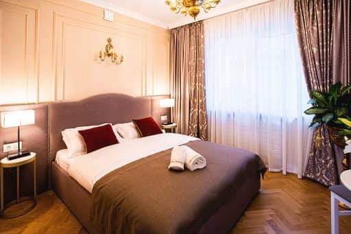 Elegancka sypialnia z zasłonami: brązowe, wzorzyste zasłony boczne i białe, prześwitujące firanki, łóżko z bordowymi poduszkami, parkiet w jodełkę.