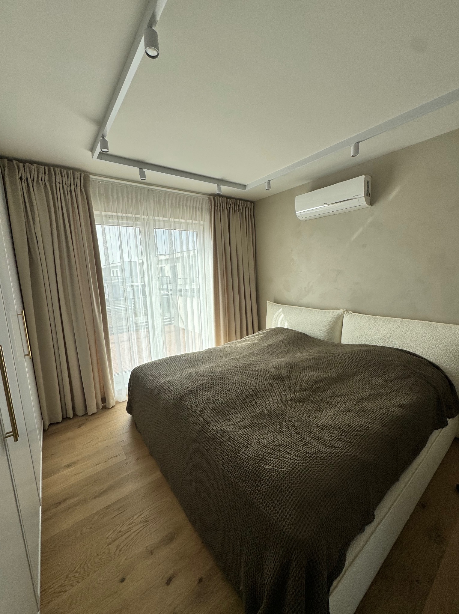 Jasna sypialnia z beżowymi, długimi zasłonami i firankami przy oknie balkonowym, brązowa narzuta na łóżku, drewniana podłoga i biała klimatyzacja na ścianie.