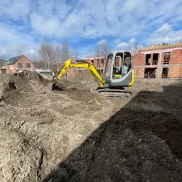 Żółto-szara mini koparka podczas prac ziemnych na placu budowy, z widocznymi fundamentami budynków w tle i błękitnym niebem z chmurami.
