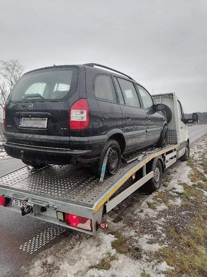 Czarny samochód osobowy Opel Zafira z uszkodzonym kołem, zabezpieczony pasami transportowymi, przewożony na platformie lawety w pochmurny dzień.
