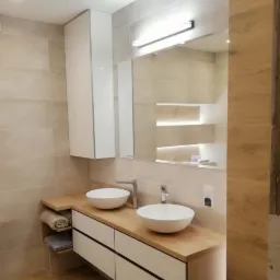 Nowoczesna łazienka z dwoma okrągłymi umywalkami nablatowymi, blatem z jasnego drewna, białymi szafkami z czarnymi uchwytami, lustrem z podświetleniem i płytkami w odcieniach beżu.