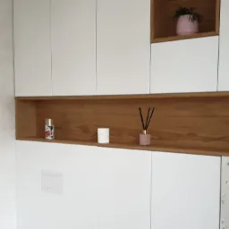 Białe szafki łazienkowe na wymiar z drewnianymi wnękami, w których znajdują się dekoracje, widoczna toaleta.