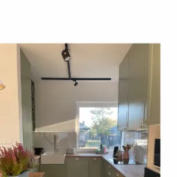 Jasna kuchnia w odcieniach zieleni z drewnianym blatem, widok na zewnątrz przez okno, podłoga ułożona w jodełkę, na blacie kuchennym ustawione akcesoria i zioła w doniczce.