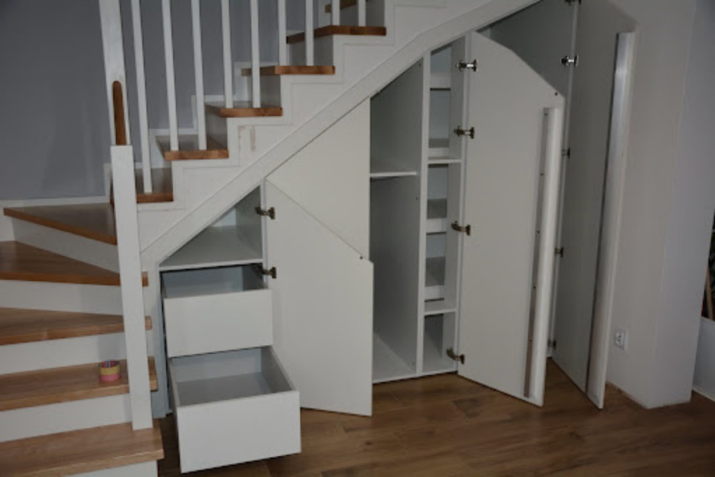 Biała zabudowa pod schodami z otwartymi szafkami i szufladami, widoczne półki wewnątrz, drewniane schody z białymi balustradami.