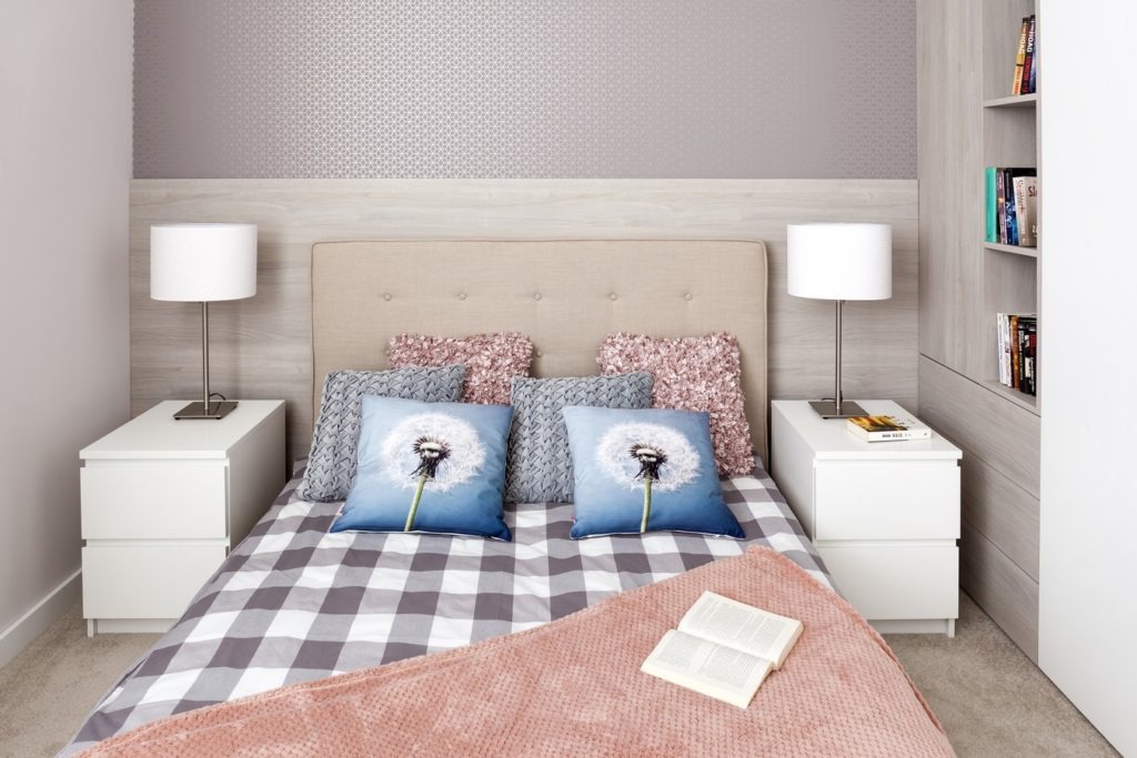 Nowoczesna sypialnia z łóżkiem, szafkami nocnymi i regałem w zabudowie w odcieniach szarości i bieli, poduszki dekoracyjne z motywem dmuchawca, książka na łóżku.