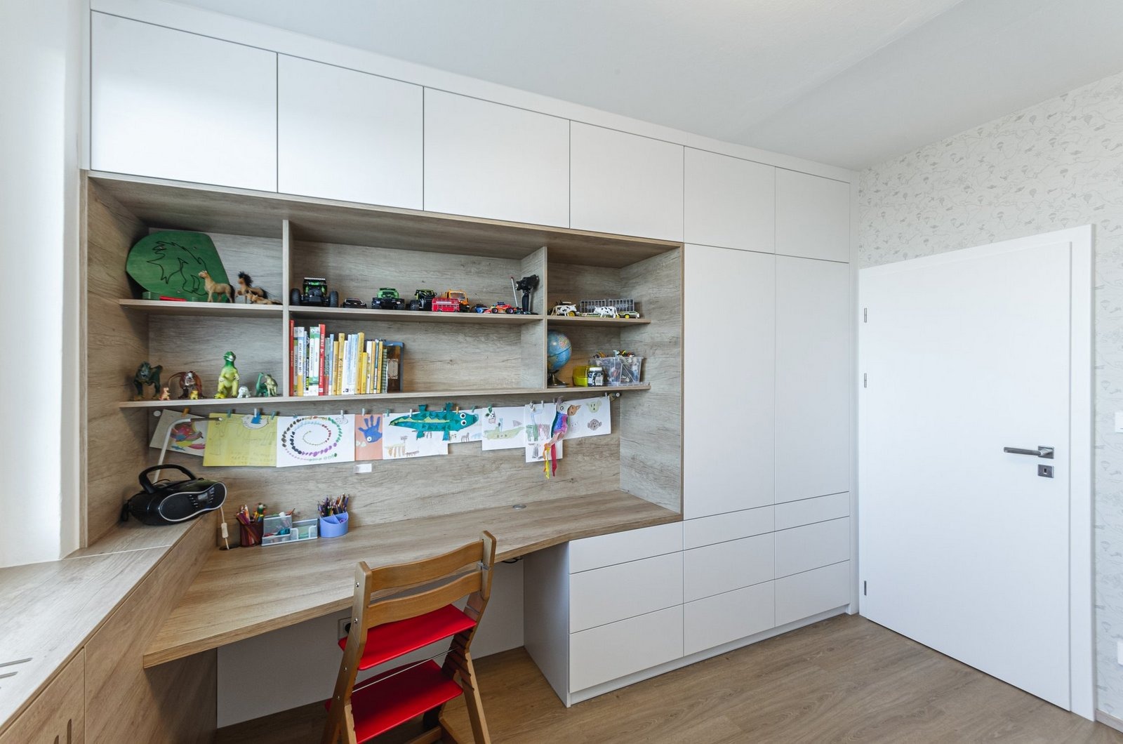 Pokój dziecięcy z biurkiem i regałem w zabudowie, białe szafki i szuflady, drewniane blaty i półki, dekoracje dziecięce na ścianie i półkach, krzesło z czerwonym siedziskiem.