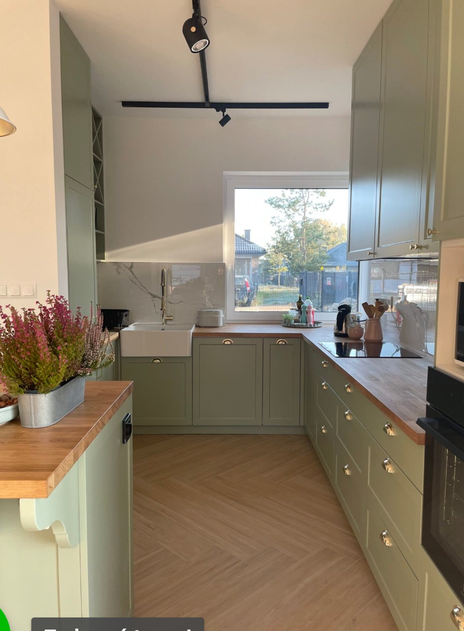 Jasna kuchnia w odcieniach zieleni z drewnianymi blatami, widok na zewnątrz przez okno, podłoga ułożona w jodełkę, oświetlenie szynowe