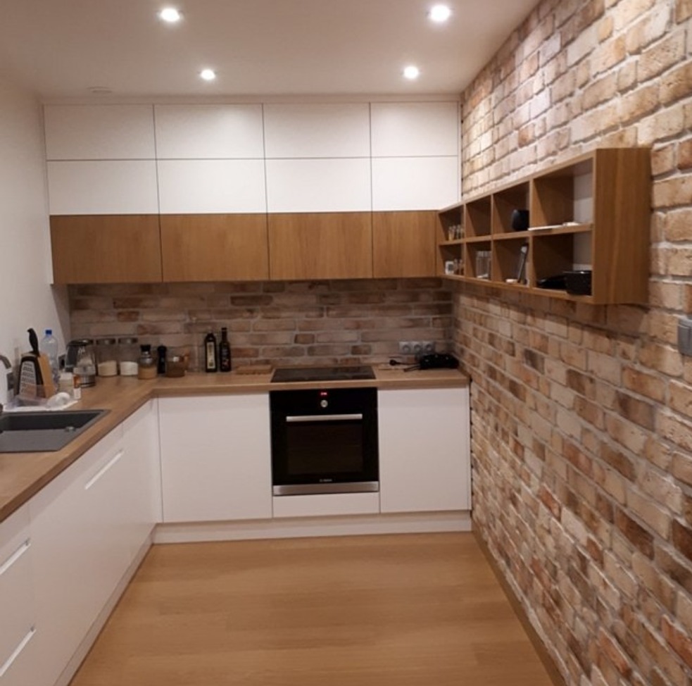 Nowoczesna kuchnia w stylu loft z białymi szafkami dolnymi i górnymi, drewnianymi blatami oraz ceglaną ścianą z drewnianymi półkami.